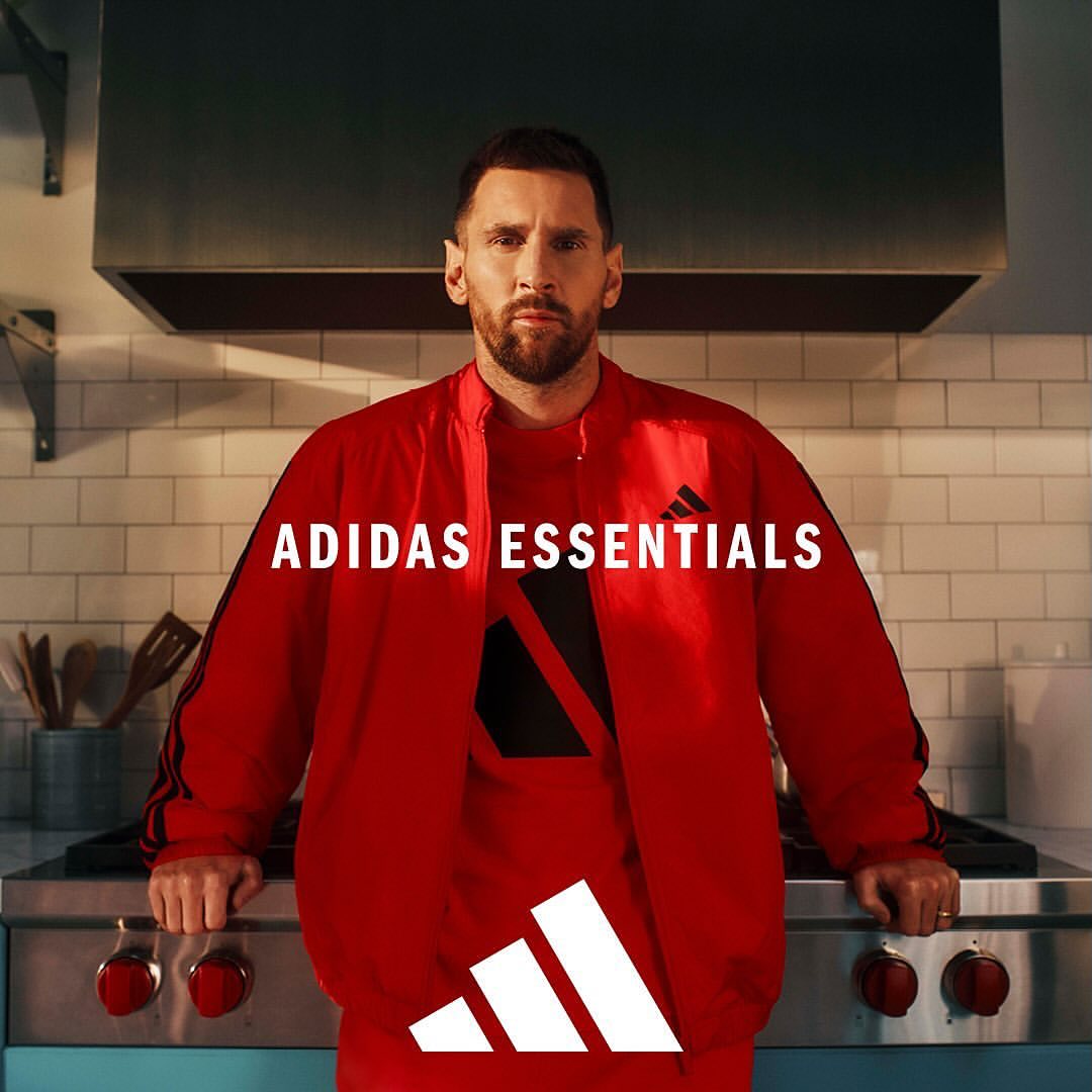 Adult man poses in red Adidas Essentials tracksuit in indoor kitchen with steel appliances and white tile backdrop and Adidas logo.