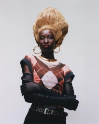 Portrait of a Black woman with large blonde sculpted hair wearing an argyle sweater, hoop earrings, and black lace gloves against a white backdrop.