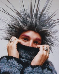 Close-up portrait of a model wearing a spiky grey textured headpiece and pulling up a dark textured sweater collar, photographed by Alex De Mora