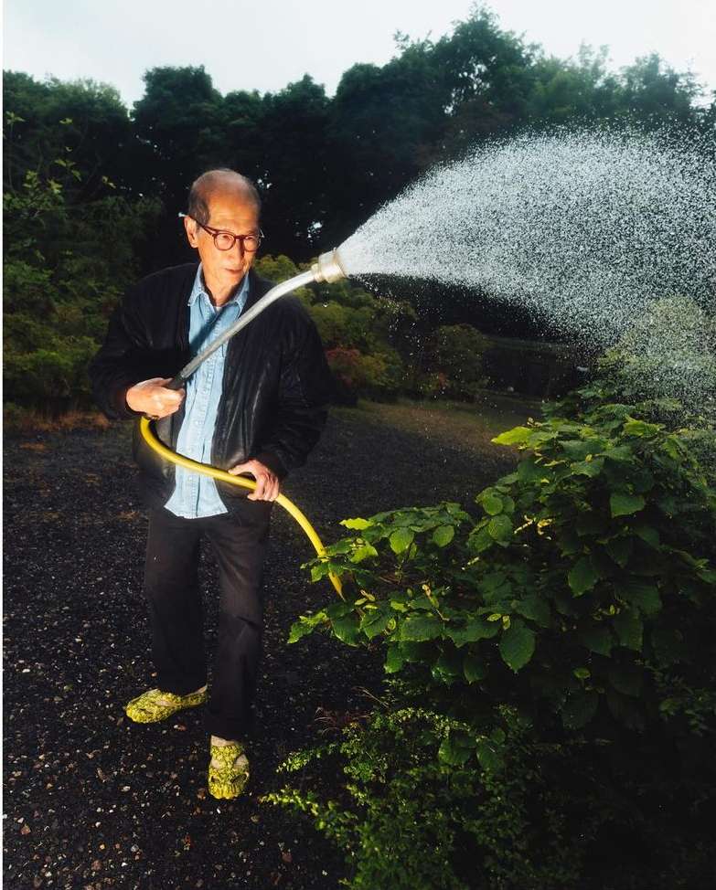 Elderly man in glasses holds yellow garden hose, watering a plant. The background shows a gravel path and green trees.