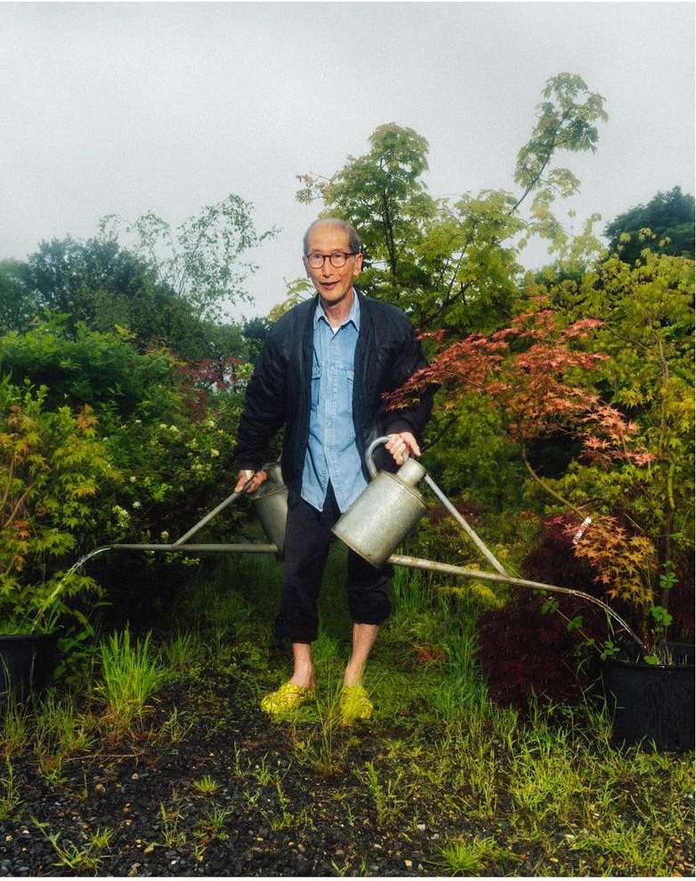 An elderly man in a garden waters plants using two watering cans, standing amongst lush greenery on a cloudy day.