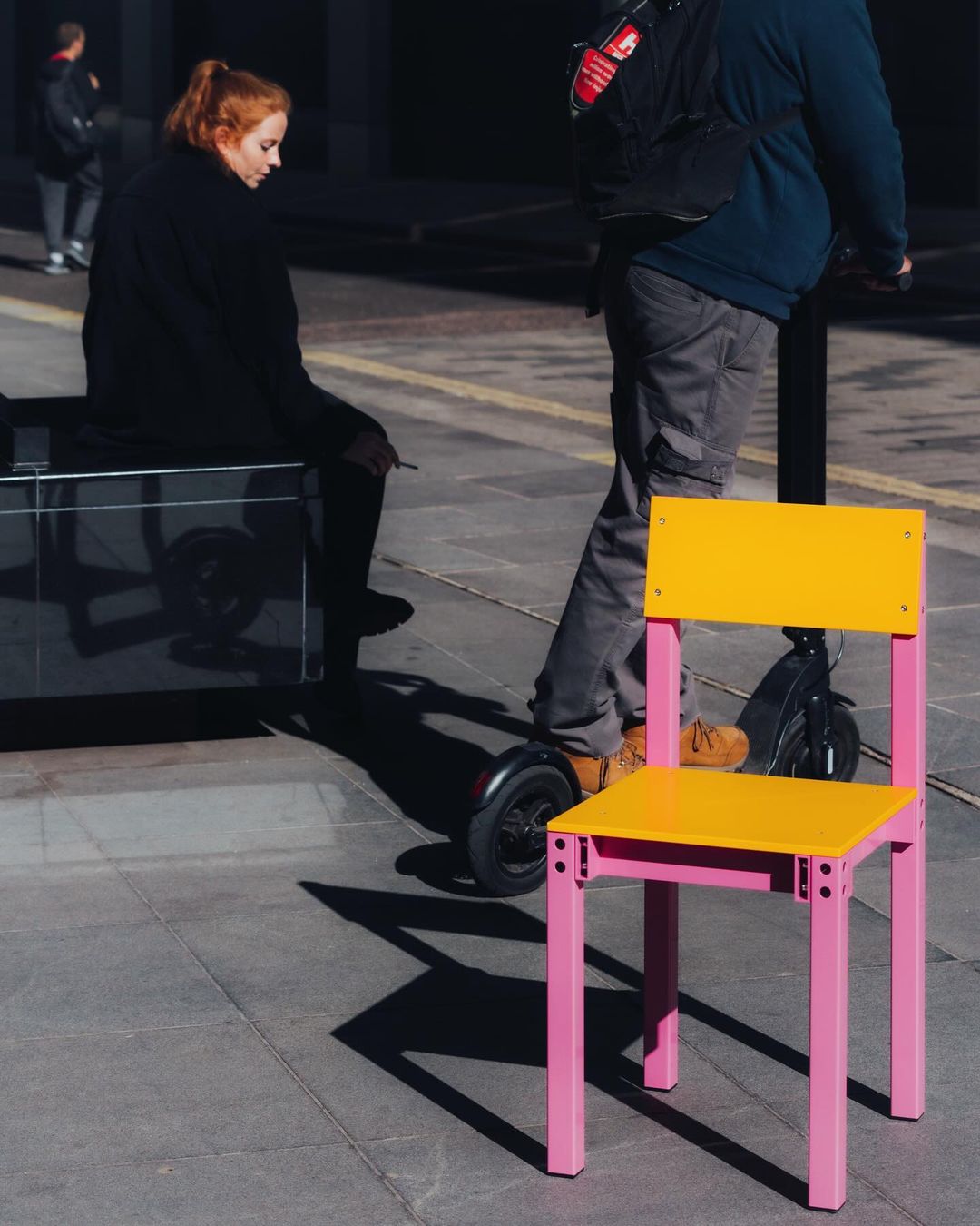 Bright yellow and pink chair sitting on an outdoor city sidewalk. Two people are walking in the background in the daylight.
