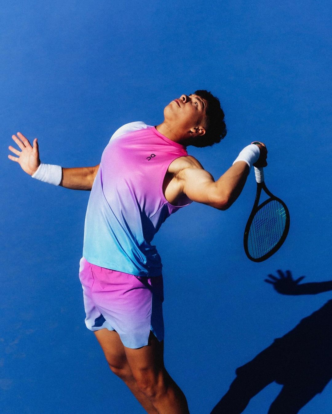Male athlete in a colorful sports uniform serves a tennis ball on a blue court with outstretched arms, ready to hit the ball with his racket.