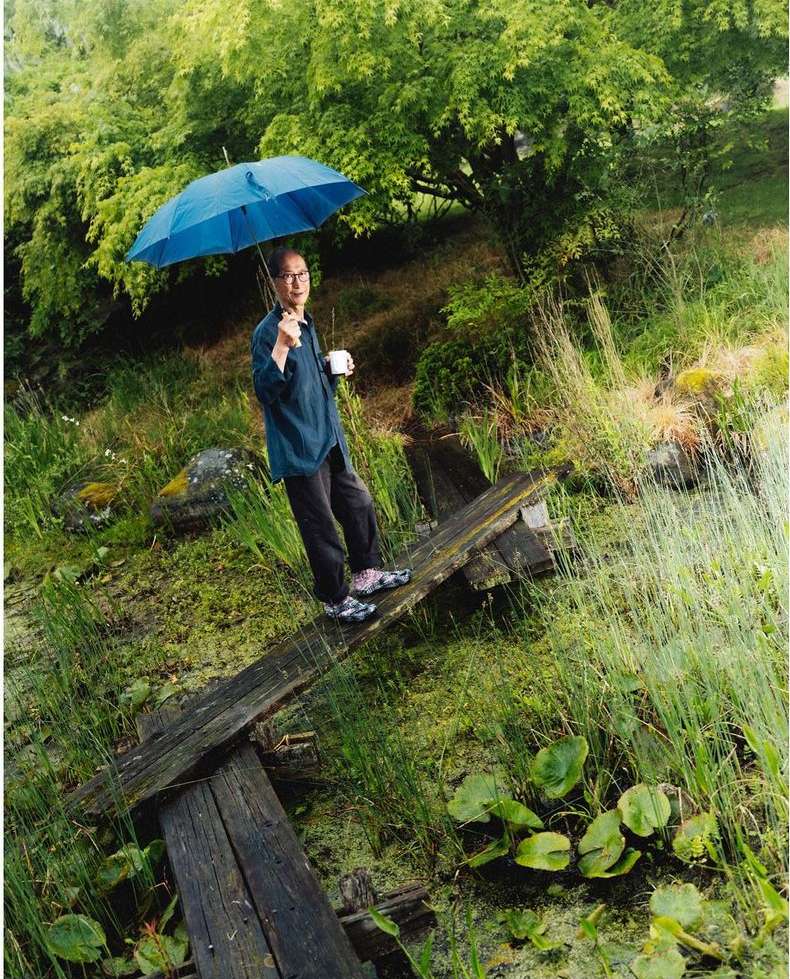 Man with glasses and an umbrella standing on a wooden plank in a marshy field with trees in the background on an overcast day.
