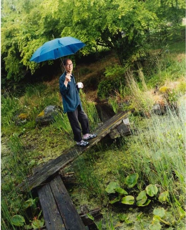 Man with glasses and an umbrella standing on a wooden plank in a marshy field with trees in the background on an overcast day.