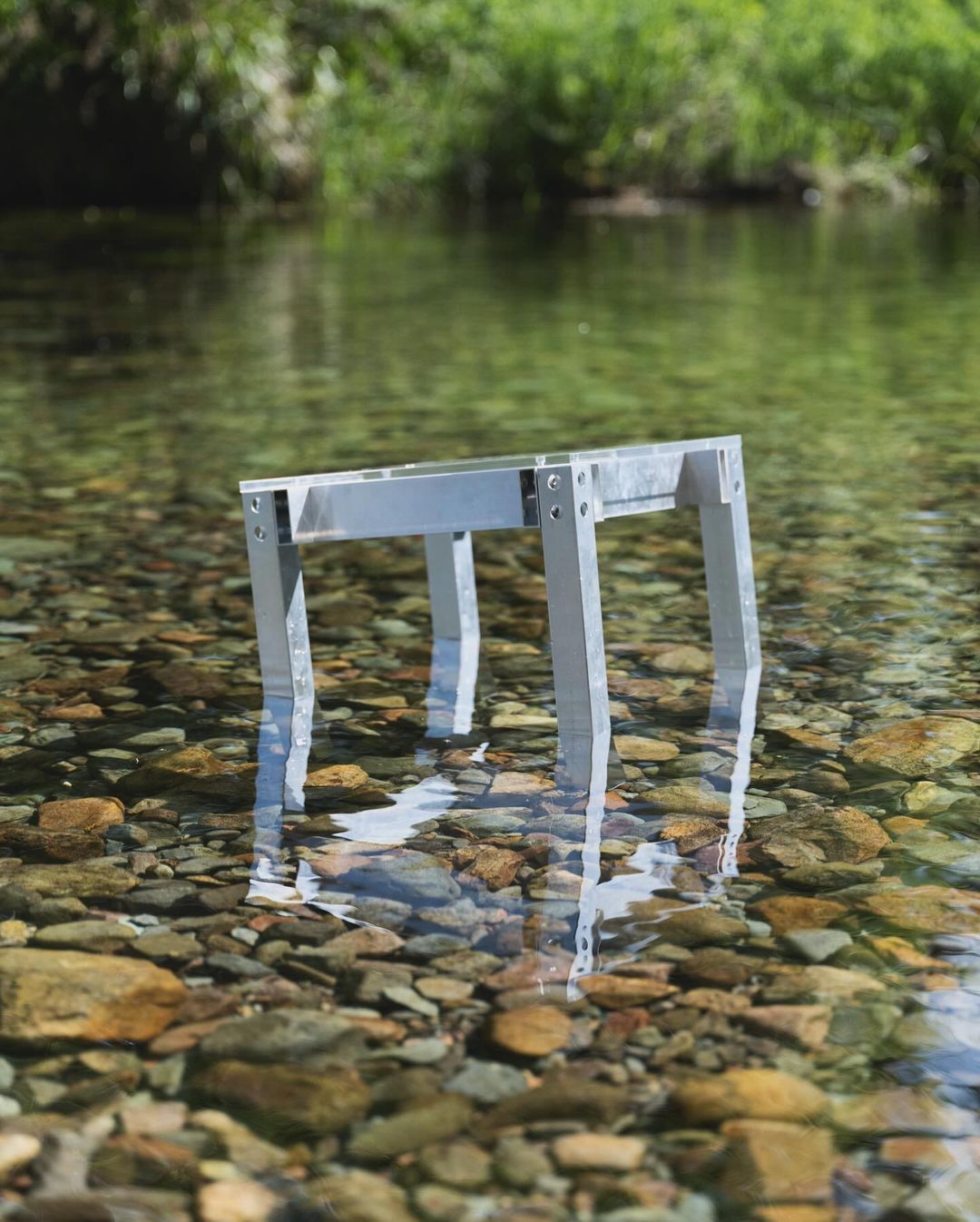 Metal structure stands in a shallow creek bed with clear water reflecting the sunlight, against a green tree-lined background.