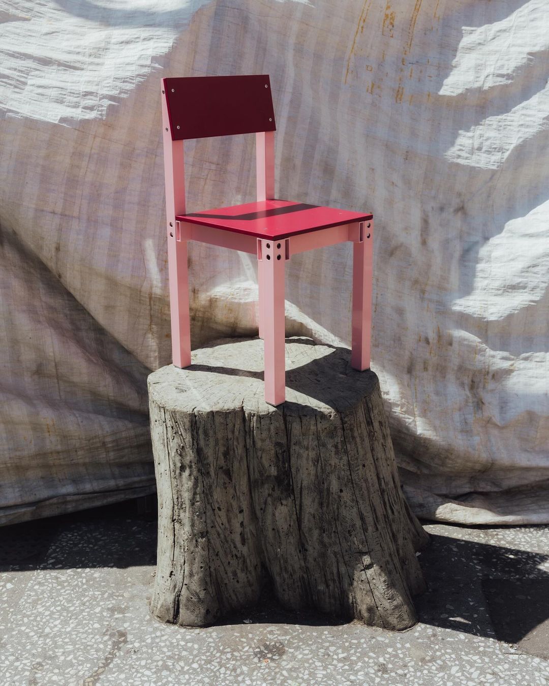 Pink and red painted wooden chair sitting on a tree stump outdoors, photographed against a backdrop of draped white fabric.