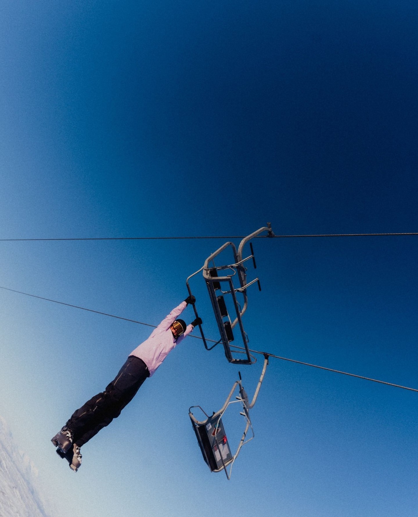 Person hanging from a ski lift, wearing ski gear, captured against a clear blue sky and snowy mountain landscape during daytime.