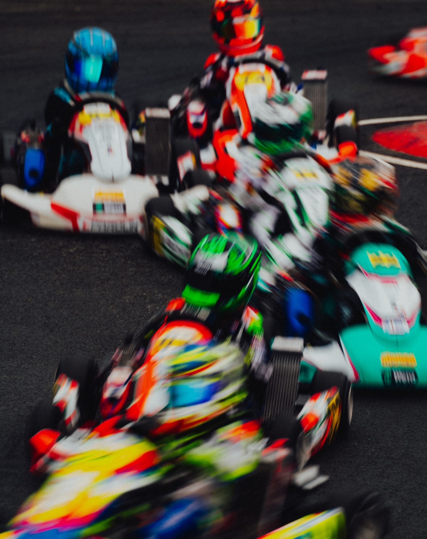 High-speed go-kart race with multiple racers in colorful helmets and racing suits competing on an asphalt track, captured with motion blur.