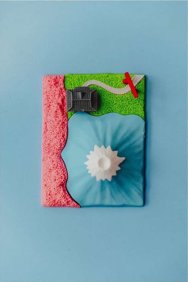 Miniature landscape art: a toy town by a lake featuring a white mountain, created with pink sand, light blue water, and green grass.