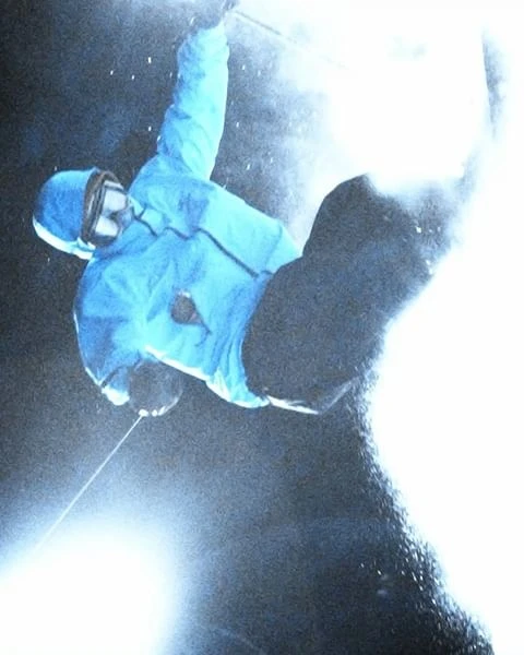 Action shot of an adult skier with a light blue jacket, black ski pants, and ski goggles performing a stunt jump at night.