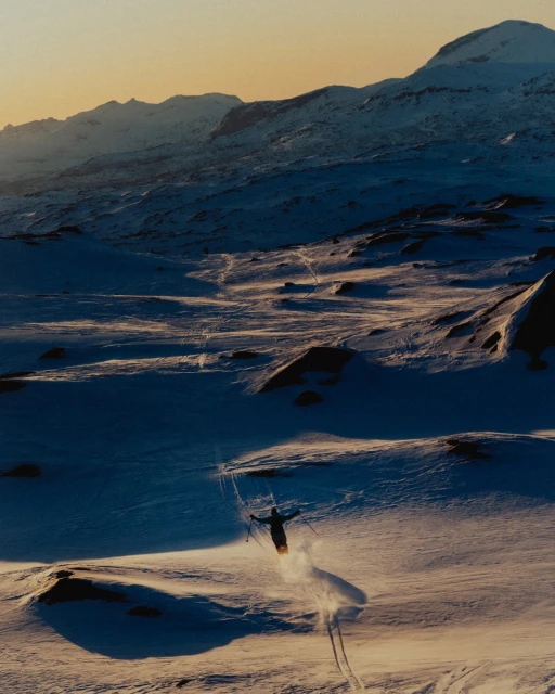 A skier is captured from above while skiing down a snowy mountain, with soft lighting and a snow-covered mountain range in the distance.