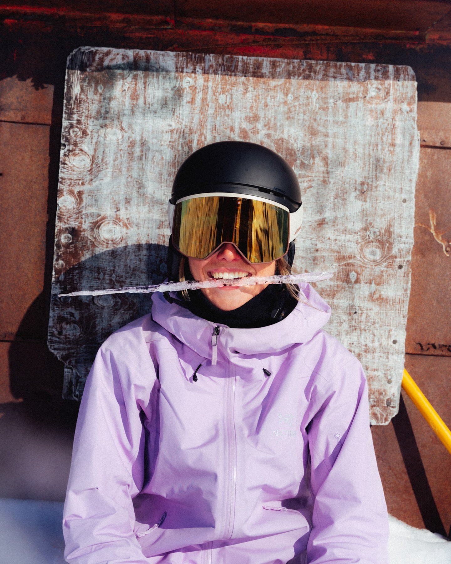 Woman in ski gear smiles with an icicle in her mouth in a snowy outdoor setting, wearing a helmet and goggles, lilac jacket.