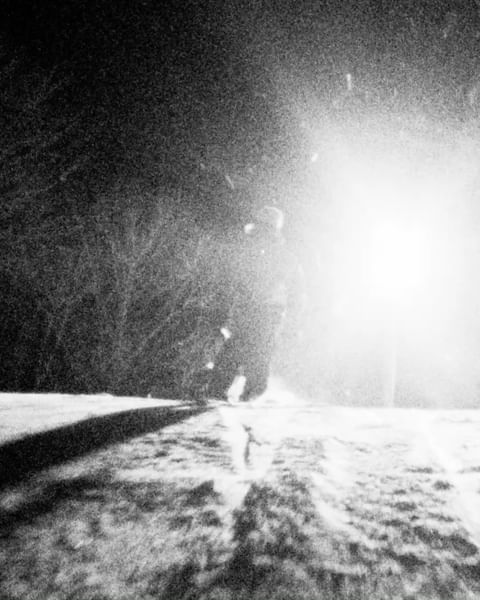 Monochromatic shot of a small figure walking on a snowy path at night with trees on the side and bright light shining in the distance.