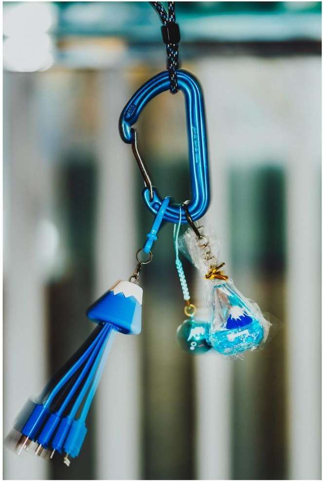 Blue carabiner attached to Mt. Fuji shaped charger cable and wrapped charm hanging against a blurred background.