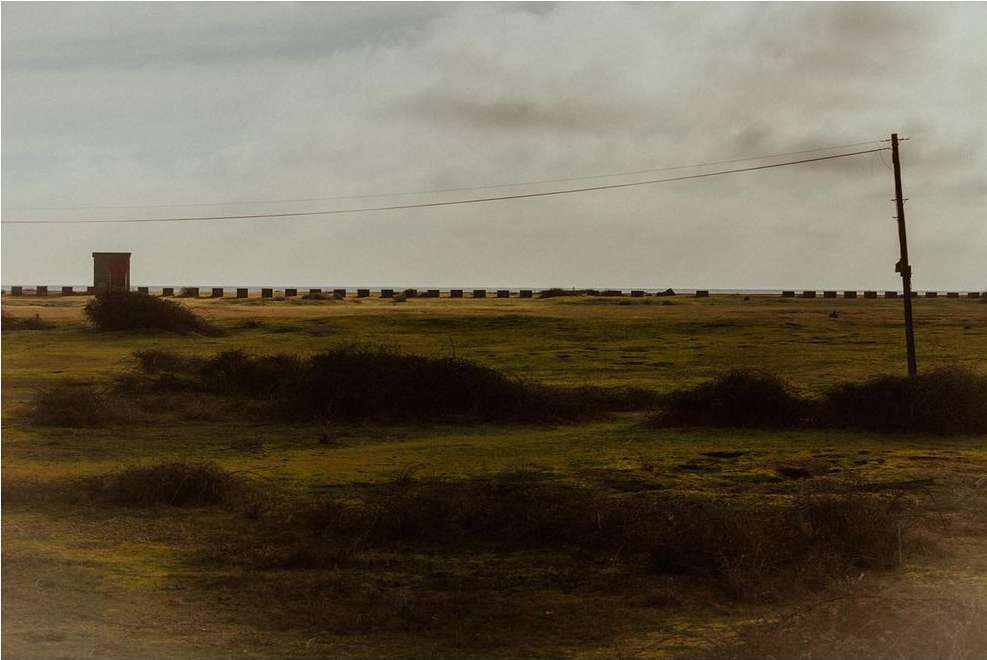 Tranquil landscape featuring a flat green field, distant building, concrete blocks, utility pole, and serene overcast sky.