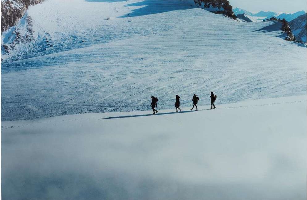Four people traverse a snowy mountain landscape with ice formations, partially obscured by a layer of low-lying clouds, creating a striking vista.