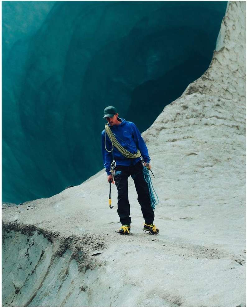 A lone climber in a blue jacket and cap stands on an icy cliff, holding an ice ax, with a blue glacier cave behind him.