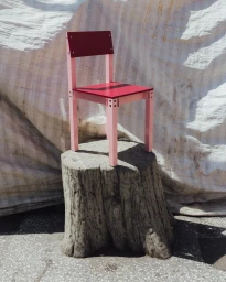 Pink and red wooden chair resting on a large tree stump against a draped white fabric backdrop outdoors.