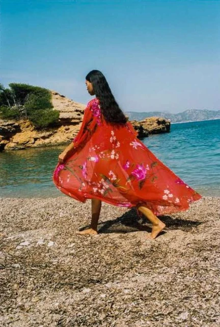Woman in red floral kimono walks on a pebbled beach near the ocean on a sunny day with a rocky cliff in the background.