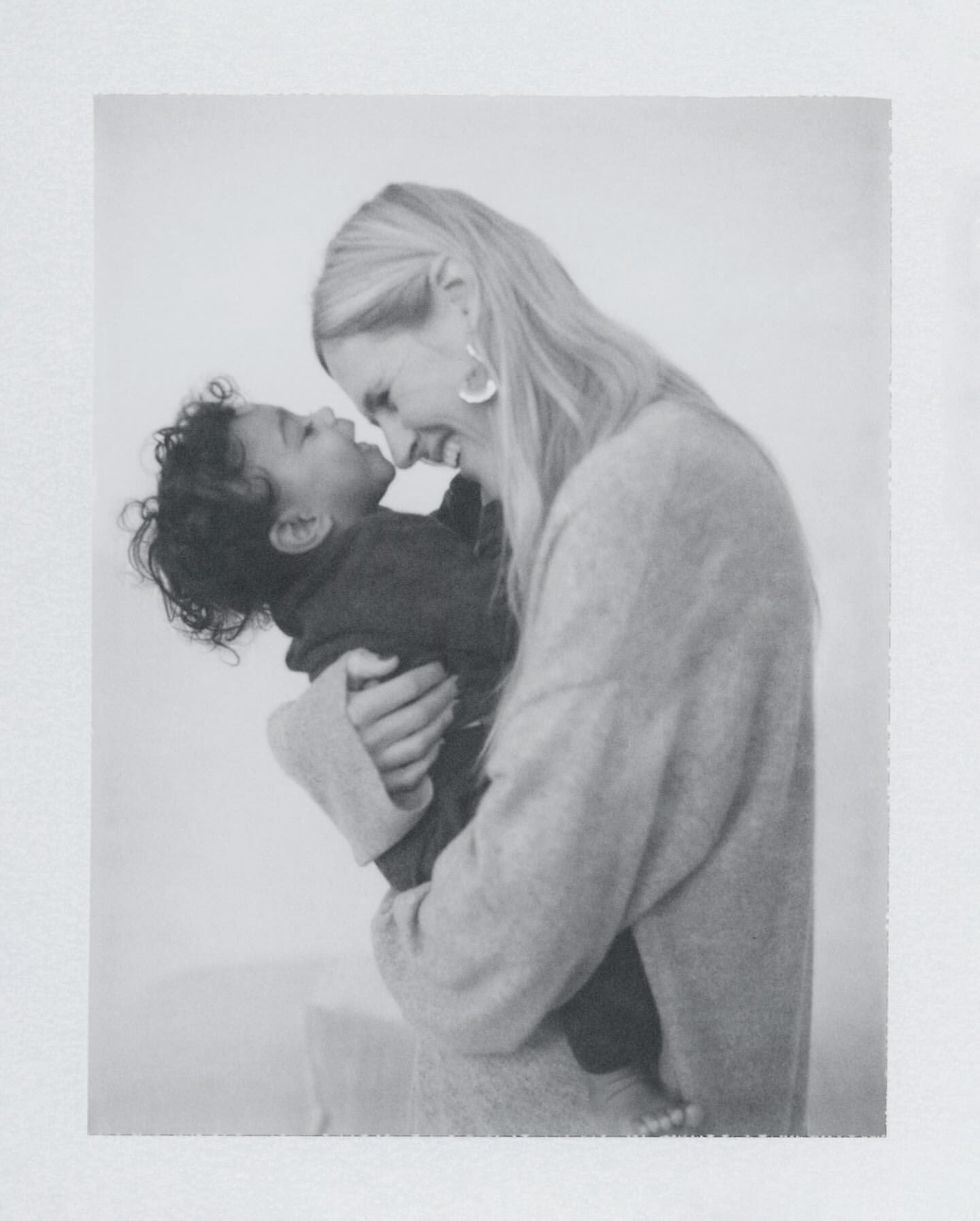 Monochrome image of a blonde adult woman lovingly holding a baby. The indoor setting features soft, diffused lighting.