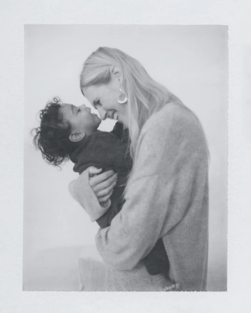 Monochrome image of a blonde adult woman lovingly holding a baby. The indoor setting features soft, diffused lighting.
