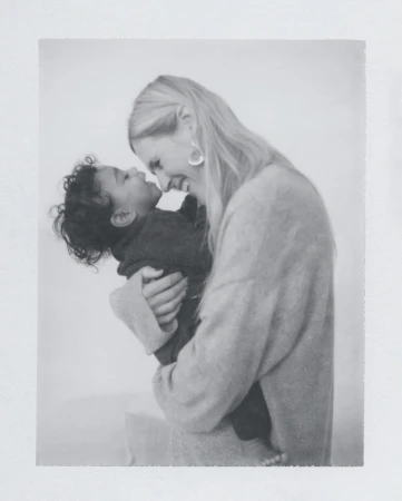 Monochrome image of a blonde adult woman lovingly holding a baby. The indoor setting features soft, diffused lighting.