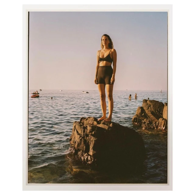 Adult woman standing on a large rock in the sea, wearing a swimsuit, with a blue sky, other people, and watercraft visible in the background.