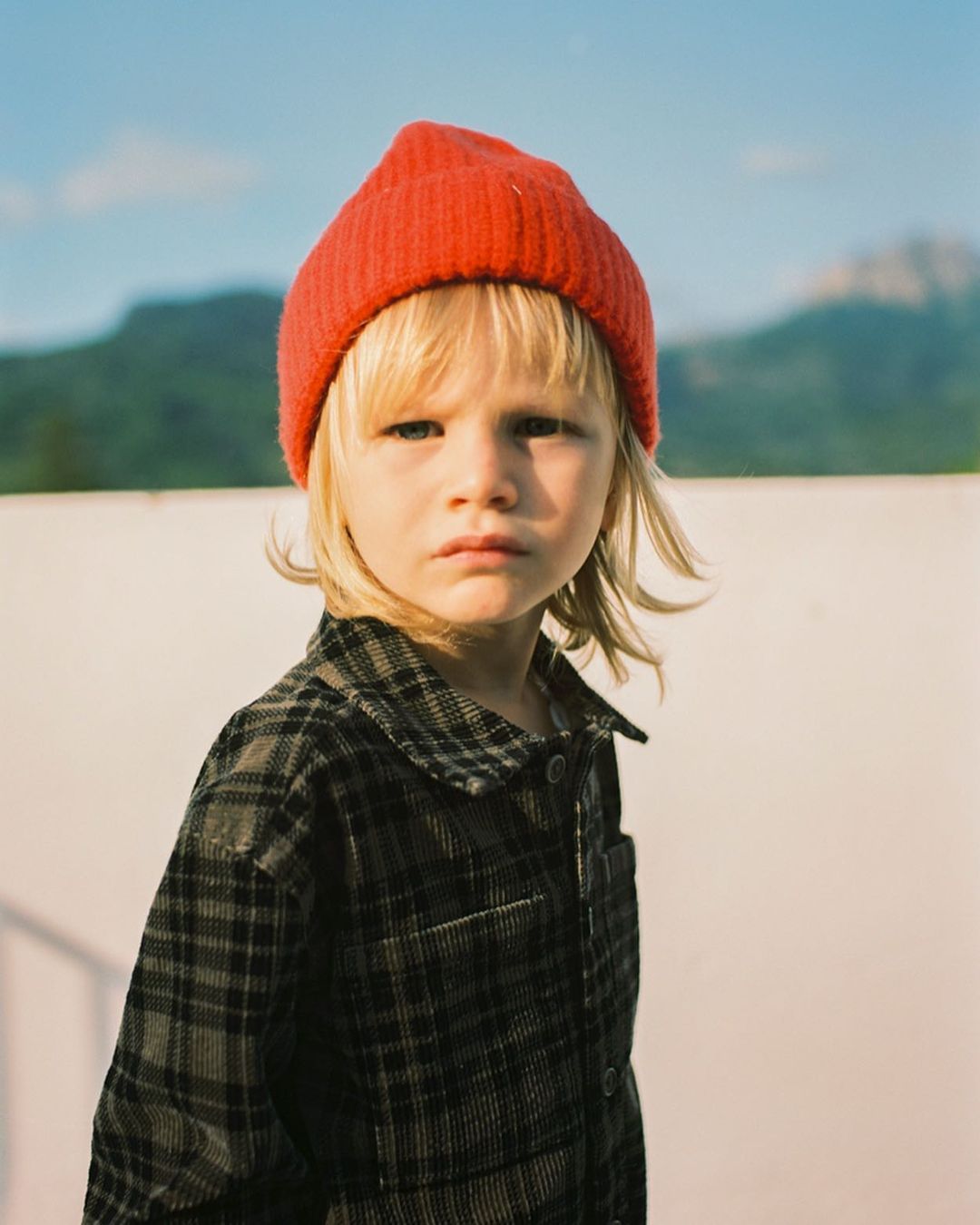 Portrait of a blonde child in a red beanie and plaid jacket posing outdoors with mountains visible in the background under a blue sky.