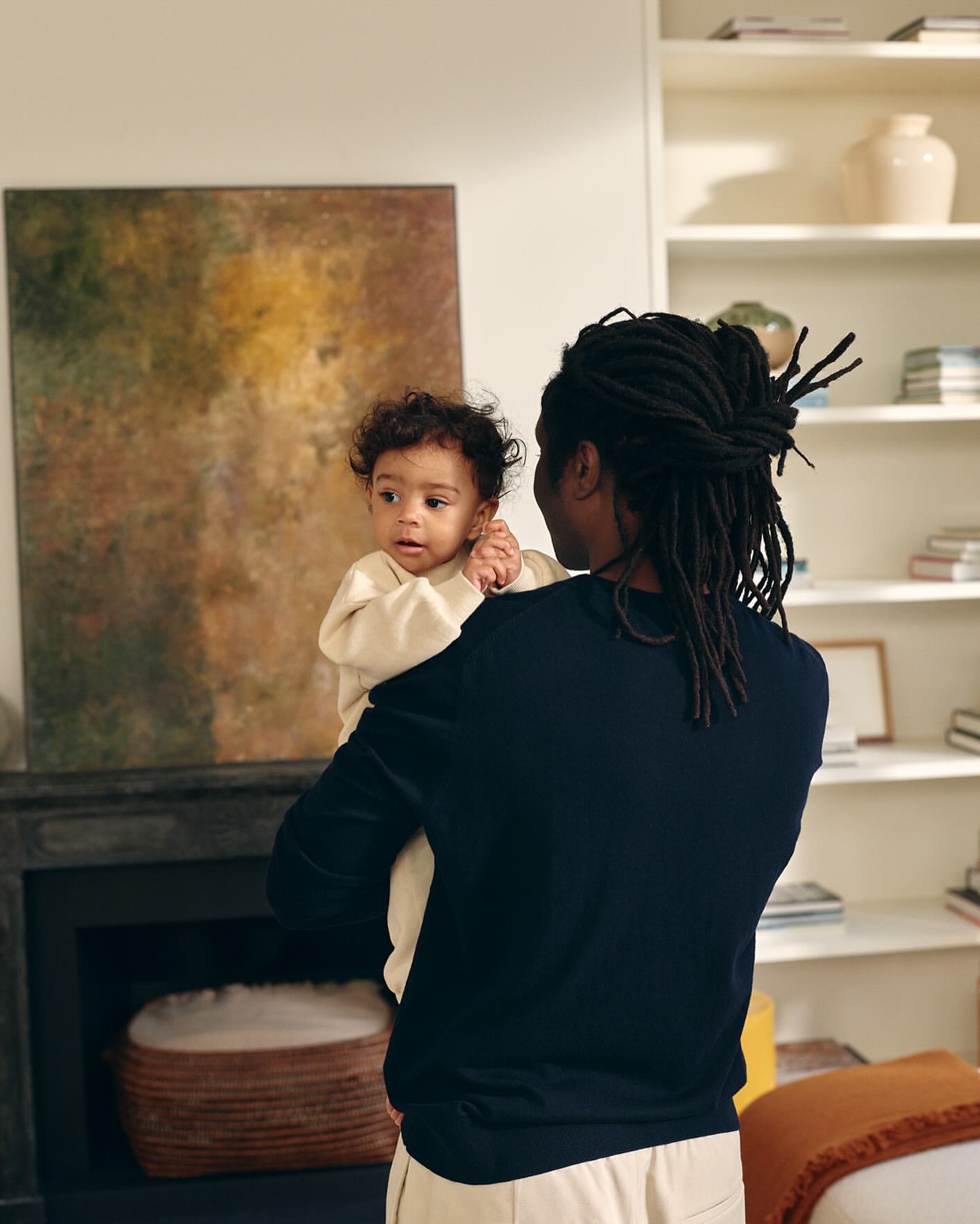 Dark-haired adult holds a young child indoors, standing near a fireplace with artwork and a white bookshelf in a softly lit room.