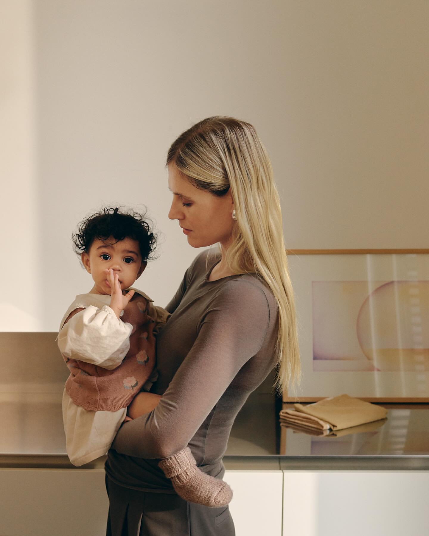 Blonde mother indoors holds her curly-haired baby near cabinetry and artwork, showcasing a tender moment and a glimpse of home interior design.