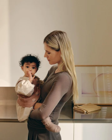 Blonde mother indoors holds her curly-haired baby near cabinetry and artwork, showcasing a tender moment and a glimpse of home interior design.
