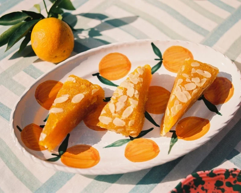 Overhead shot of orange desserts topped with almonds on a decorative plate with painted oranges and leaves, with fresh orange.