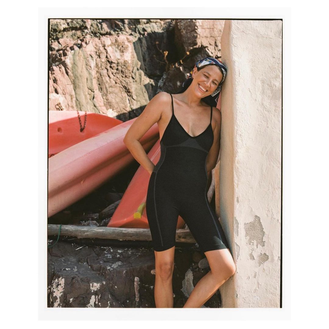 Woman in black activewear and headscarf smiles outdoors against a sunlit wall, with stacked kayaks and rock formations in the background.