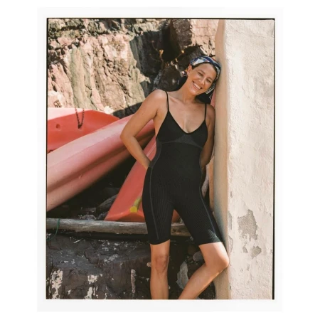 Woman in black activewear and headscarf smiles outdoors against a sunlit wall, with stacked kayaks and rock formations in the background.