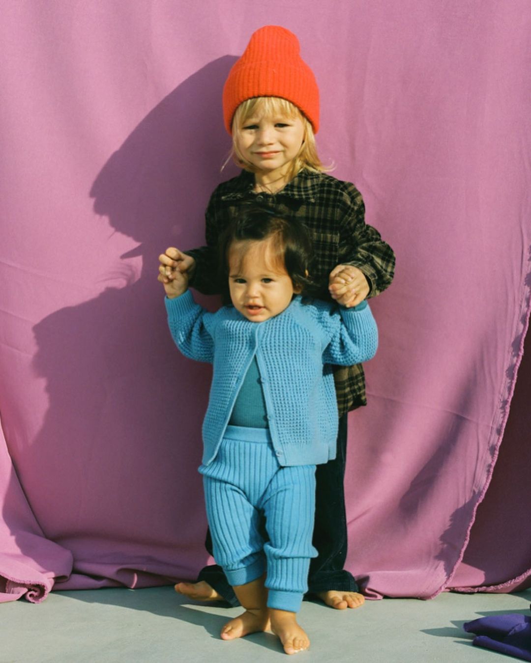 Two young children, a toddler in a blue knit outfit and an older child in a plaid shirt and orange beanie, pose in front of a lilac backdrop.