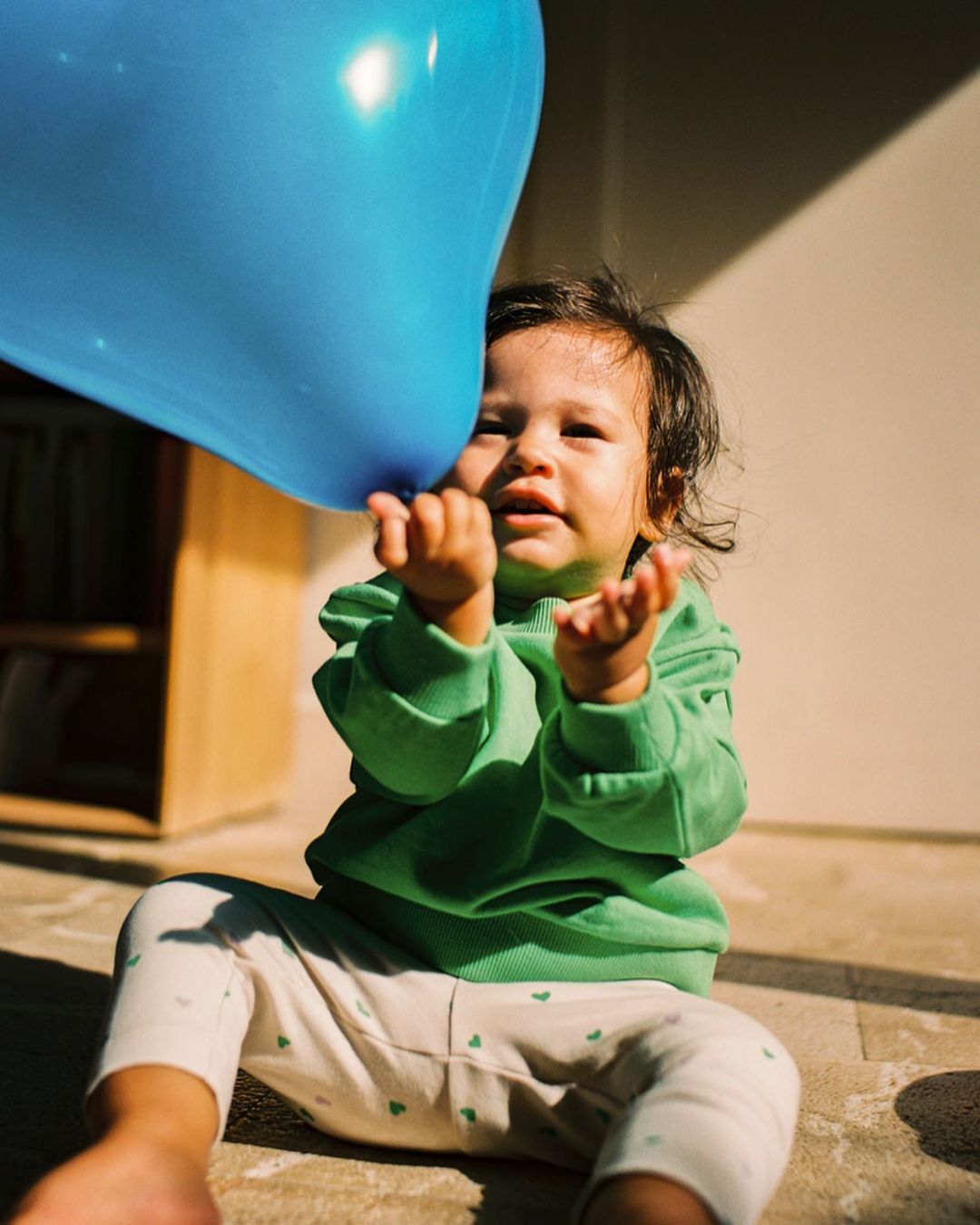 A child sitting outdoors reaches for a large blue balloon, bathed in warm sunlight, wearing a green sweatshirt and patterned leggings.