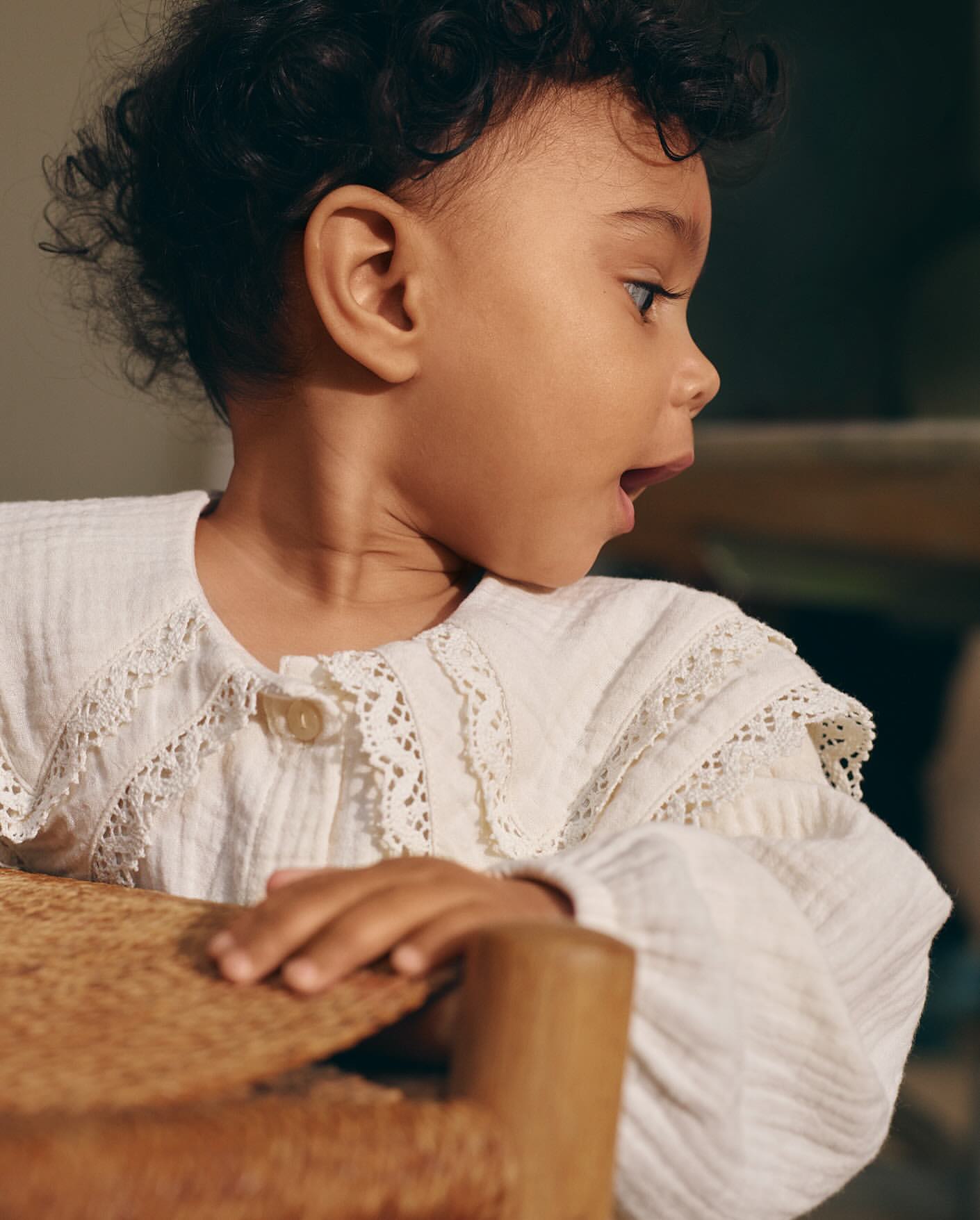A young child with curly hair, wearing a white blouse with lace trim, looks to the side with a slightly open mouth in a close-up shot.