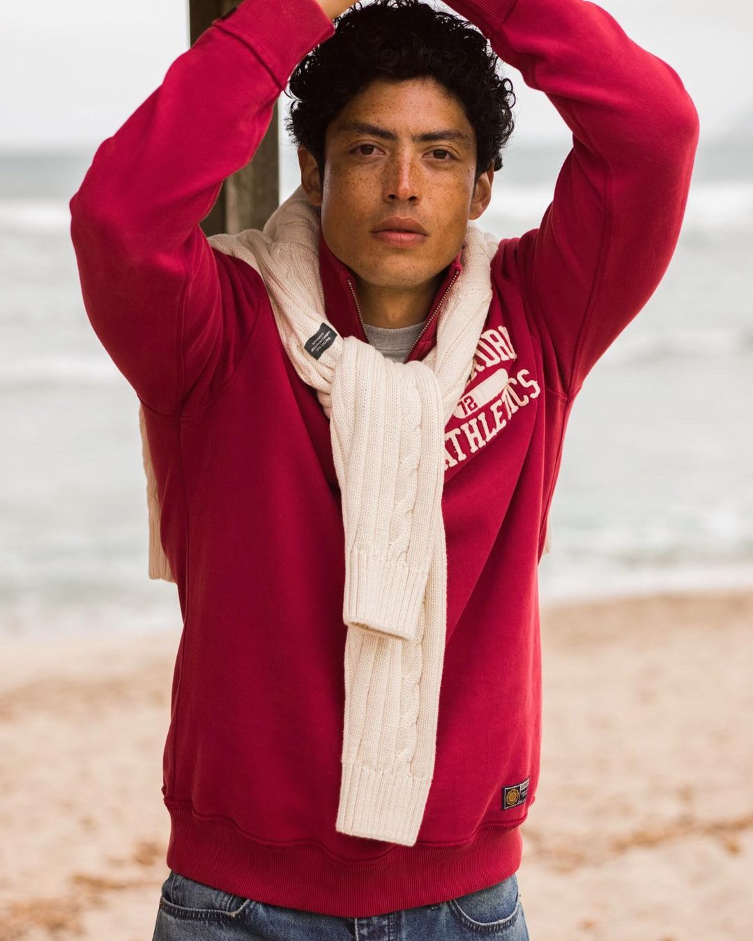 An adult male with dark curly hair wearing a red sweatshirt and jeans stands on a sandy beach with ocean in the background.