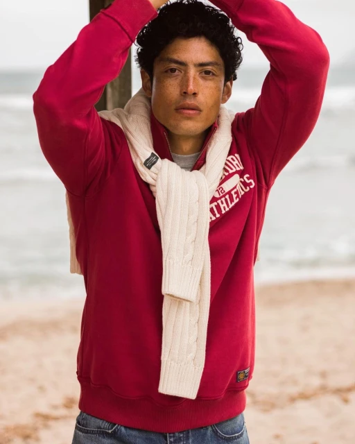 An adult male with dark curly hair wearing a red sweatshirt and jeans stands on a sandy beach with ocean in the background.