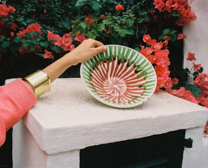 A person holds a decorative plate with red and green spirals against a background of red flowers and white stucco.