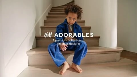 Young boy with curly hair poses barefoot in blue outfit on wood stairs for H&M Adorables campaign; neutral colors, warm lighting.
