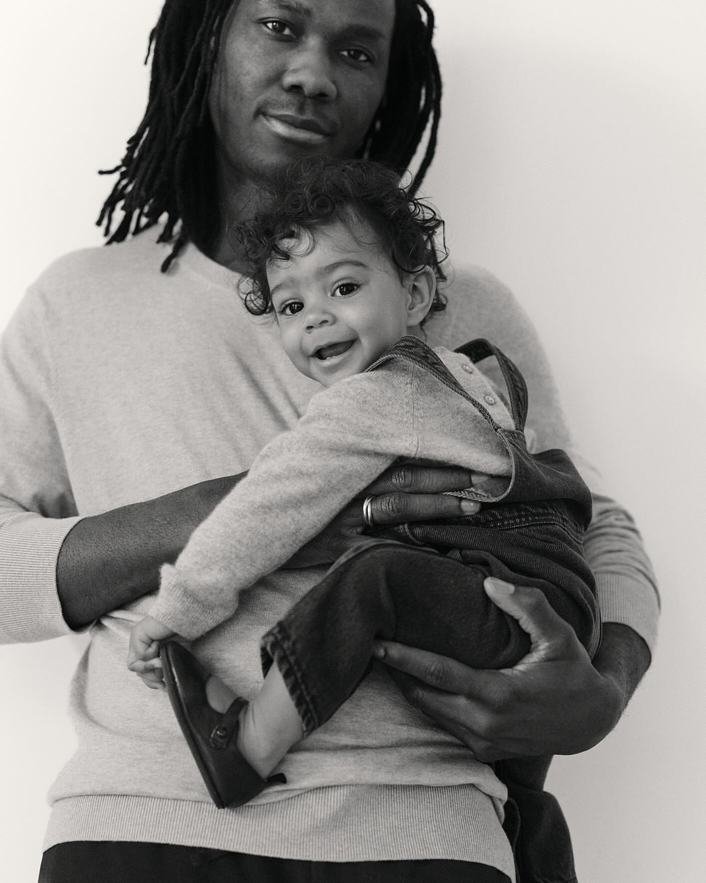 Black and white portrait of a Black adult male holding a happy baby, both wearing casual sweaters, against a plain background.