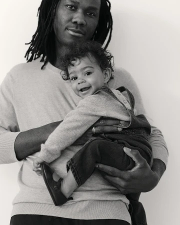 Black and white portrait of a Black adult male holding a happy baby, both wearing casual sweaters, against a plain background. 