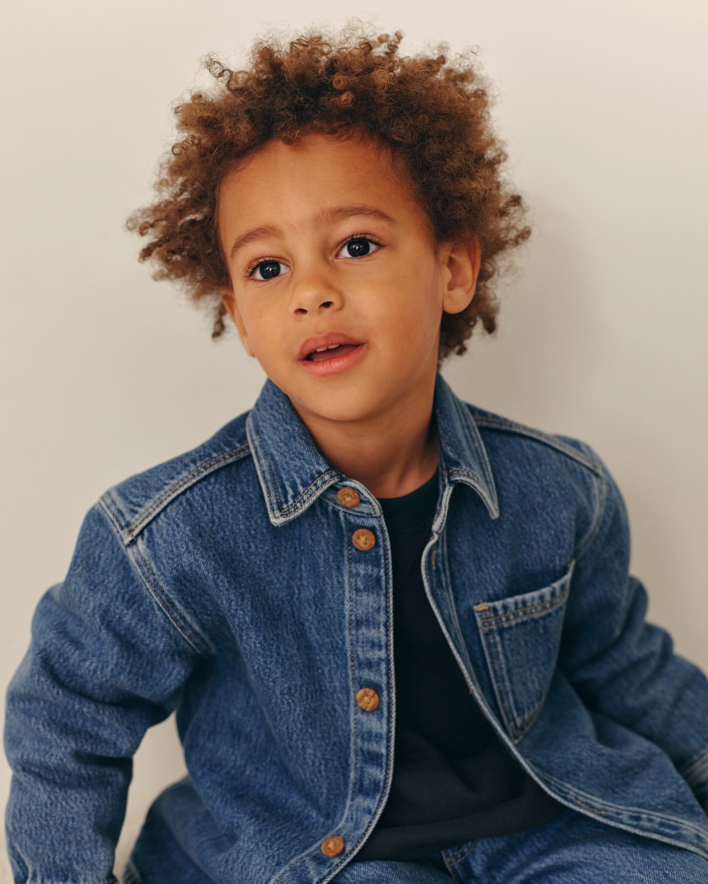 Young child with curly brown hair wearing a denim shirt against a plain background