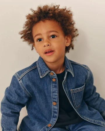 Young child with curly brown hair wearing a denim shirt against a plain background