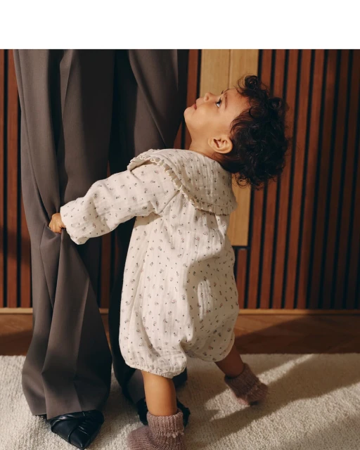 A baby in a floral romper stands next to an adult in dark trousers, creating a heartwarming contrast of innocence and sophistication.