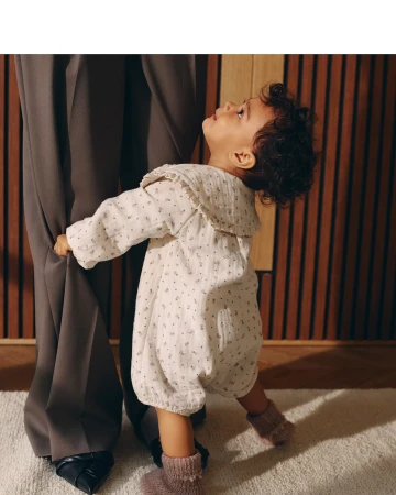 A baby in a floral romper stands next to an adult in dark trousers, creating a heartwarming contrast of innocence and sophistication.