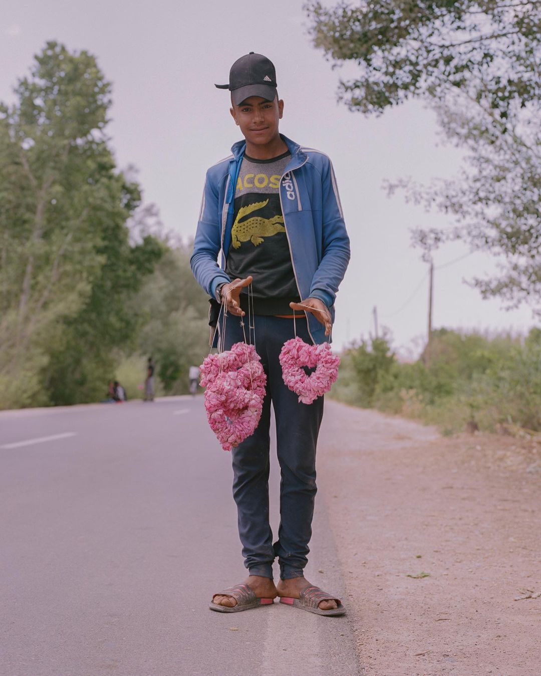 Young adult stands on a road with heart-shaped floral arrangements, trees and sky visible in the background on an overcast day.
