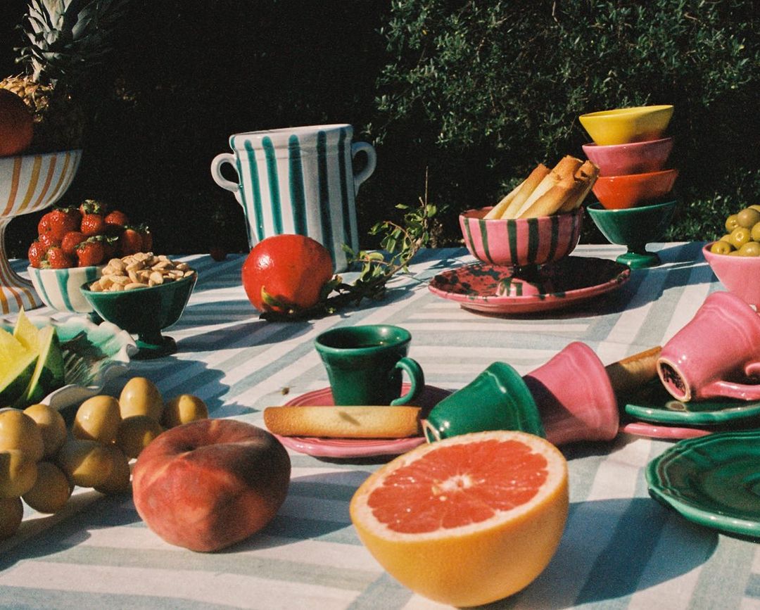 A table laden with colorful dishware and fresh fruit, including strawberries, grapefruit, and peaches, set outdoors in natural sunlight.